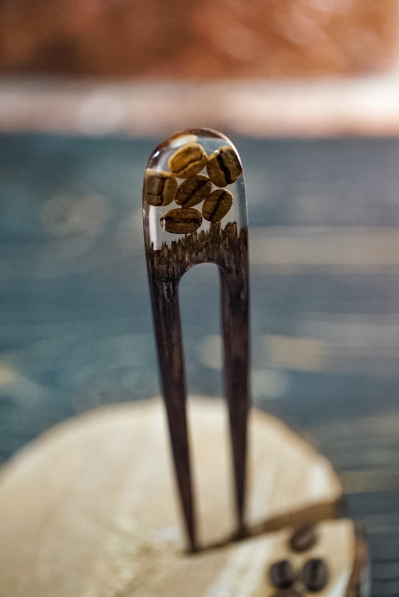 May include: A wooden hair fork with a clear resin top containing coffee beans. The fork is resting on a wooden surface.