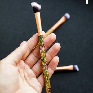 May include: Makeup brush with a gold ferrule and a handle filled with dried flowers. The brush has a white and black angled brush head. Two additional brushes are slightly out of focus in the background.