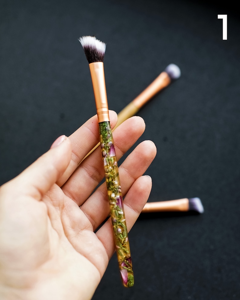 May include: Makeup brush with a gold ferrule and a handle filled with dried flowers. The brush has a white and black angled brush head. Two additional brushes are slightly out of focus in the background.