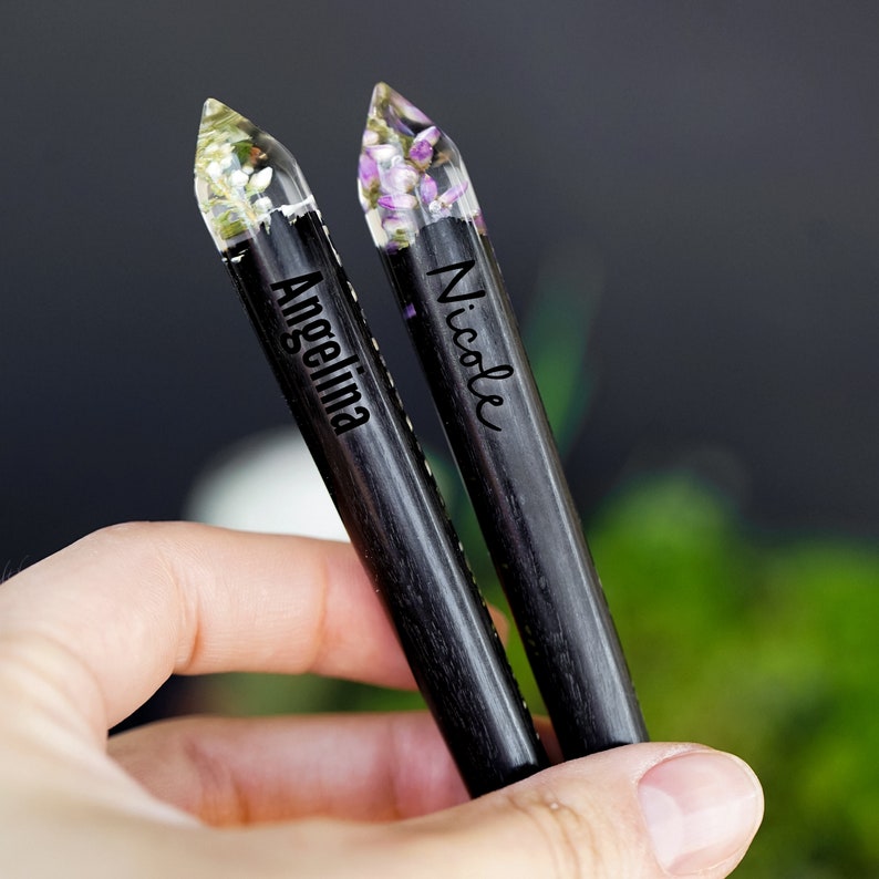 May include: Two black wooden pens with clear resin tops filled with dried flowers. The pens are engraved with the names "Angelina" and "Nicole".