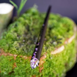 May include: A black wooden hair stick with a clear resin tip containing purple flowers. The stick is resting on a bed of green moss.
