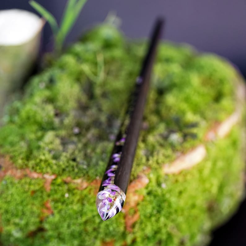 May include: A black wooden hair stick with a clear resin tip containing purple flowers. The stick is resting on a bed of green moss.