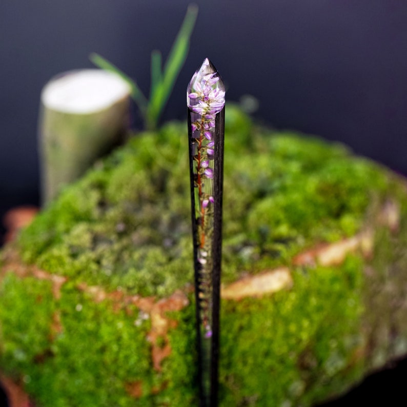 May include: A clear glass rod with a pointed tip, containing a preserved flower with pink petals. The rod is standing upright on a bed of green moss.