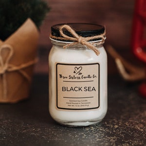 May include: A glass jar candle with a black lid and a brown paper label. The label says "Three Sisters Candle Co." and "Black Sea". The candle is scented soy and hand poured in Tennessee. It weighs 12 ounces.