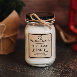 May include: A white candle jar with a brown label that reads "Three Sisters Candle Co. Christmas Hearth Scented Soy Candle Hand Poured in Tennessee Net Wt. 12 oz. (340.19 g)"