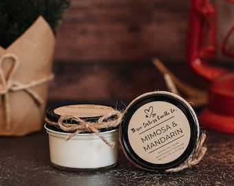 May include: Two small glass jars with lids containing soy candles. The candle jars are tied with twine and a bow. The jar on the right has a black label with white text that reads "Three Sisters Candle Co. Mimosa & Mandarin Scented Soy Candle Hand Poured in Tennessee Net Wt. 2.15 oz (77 g)"