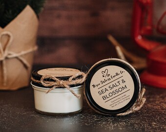 May include: Two small, white soy wax candles in glass jars with black lids, labelled "Sea Salt & Blossom" by Three Sisters Candle Co. Each jar is wrapped with twine. A small decorative tree and a red lantern are in the background.