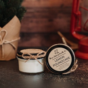 May include: Two small glass jars with lids. The jar on the left is filled with a white candle. The jar on the right has a black label with white text that reads "Three Sisters Candle Co. Lemon Poundcake Scented Soy Candle Hand Poured in Tennessee Net Wt. 2.75 oz (77.5g)"
