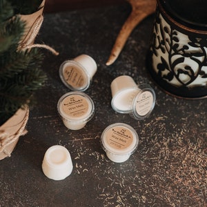 May include: Five small white wax melts in clear plastic containers with brown labels that say "Pure Serenity Candle Co. Wax Melt". The wax melts are on a dark brown textured surface with a black and gold decorative candle warmer in the background.