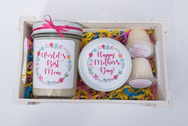 Op de afbeelding: Een witte houten doos gevuld met kleurrijk tissuepapier en drie kaarsen. De kaarsen zitten in glazen potten met bloemmotieven en de tekst "World's Best Mom" en "Happy Mother's Day!" De kaarsen zijn geparfumeerd met citroencake en katoen.