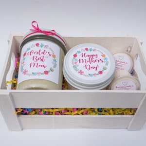 Op de afbeelding: Een witte houten kist gevuld met kleurrijk versnipperd papier en drie kaarsen. De kaarsen zitten in glazen potten met bloemenmotieven en de tekst "World's Best Mom" en "Happy Mother's Day!" De kleinere kaarsen zijn gelabeld met "Lemon Cupcake".