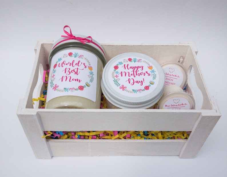 Op de afbeelding: Een witte houten kist gevuld met kleurrijk versnipperd papier en drie kaarsen. De kaarsen zitten in glazen potten met bloemenmotieven en de tekst "World's Best Mom" en "Happy Mother's Day!" De kleinere kaarsen zijn gelabeld met "Lemon Cupcake".