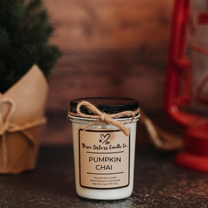May include: A small glass jar candle with a brown label that reads "Three Sisters Candle Co. Pumpkin Chai Scented Soy Candle Hand Poured in Tennessee Net Wt. 6 oz. (170.10 g)"