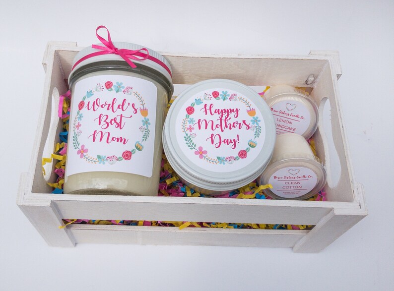 Op de afbeelding: Een witte houten kist gevuld met kleurrijk versnipperd papier en drie kaarsen. De kaarsen zitten in glazen potten met etiketten waarop staat "World's Best Mom", "Happy Mother's Day!" en "Lemon Cupcake" en "Clean Cotton".