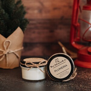 May include: Two small glass jars with lids containing soy candles. The candle on the right has a label that reads "Three Sisters Candle Co.  Plumeria Scented Soy Candle Hand Poured in Tennessee Net Wt. 2.15 oz / 77.56 g".