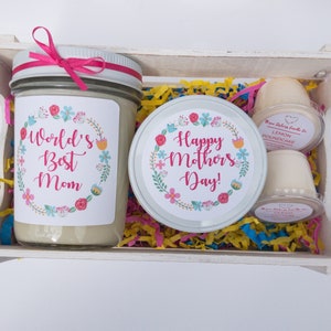 Op de afbeelding: Een witte houten kist gevuld met een cadeauset voor Moederdag. De set bevat een kaars met een bloemenmotief en de tekst "World's Best Mom", een kaars met een bloemenmotief en de tekst "Happy Mother's Day!" en twee kleine potjes met geurkaars.