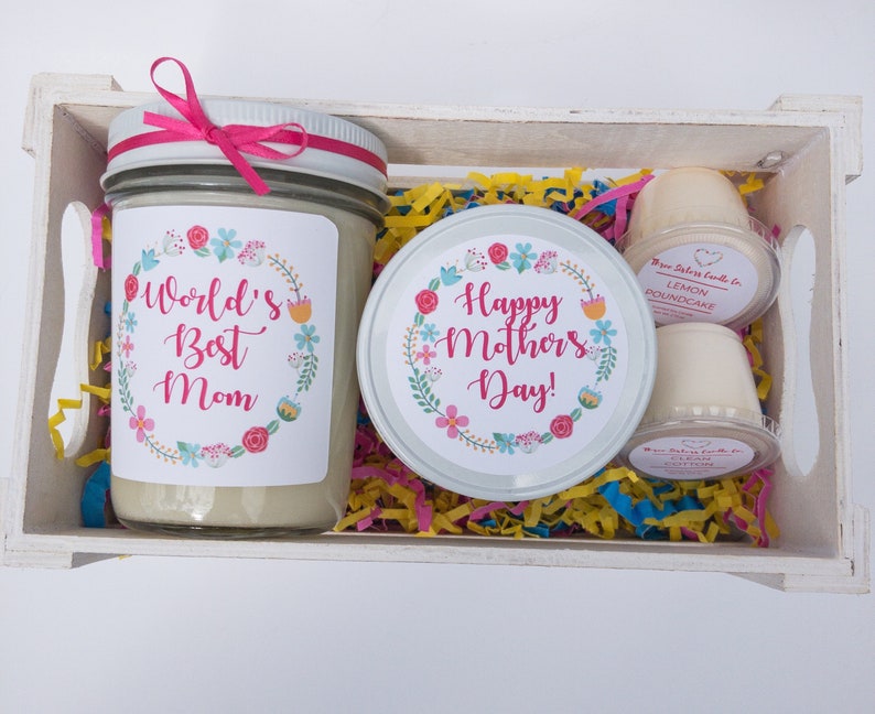 Op de afbeelding: Een witte houten kist gevuld met een cadeauset voor Moederdag. De set bevat een kaars met een bloemenmotief en de tekst "World's Best Mom", een kaars met een bloemenmotief en de tekst "Happy Mother's Day!" en twee kleine potjes met geurkaars.