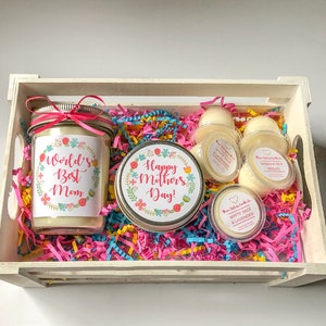 Op de afbeelding: Een witte houten kist gevuld met roze, blauwe en gele geribbeld papier. De kist bevat een kaars in een pot met een label waarop staat "World's Best Mom", een kaars in een pot met een label waarop staat "Happy Mother's Day!" en vier kleine wax melts met labels waarop staat "White Sage & Lavender" en "Banana Nut Bread".