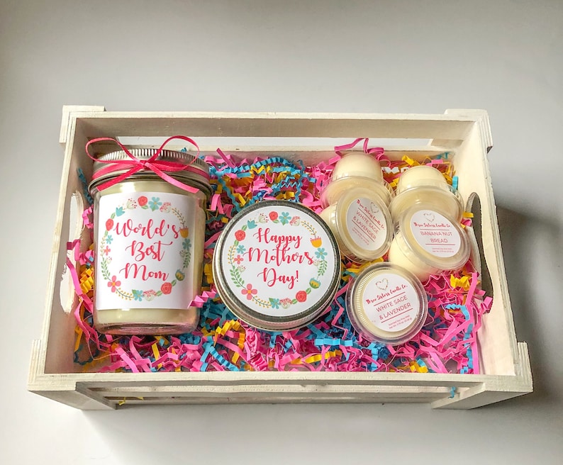 Op de afbeelding: Een witte houten kist gevuld met roze, blauwe en gele geribbeld papier. De kist bevat een kaars in een pot met een label waarop staat "World's Best Mom", een kaars in een pot met een label waarop staat "Happy Mother's Day!" en vier kleine wax melts met labels waarop staat "White Sage & Lavender" en "Banana Nut Bread".