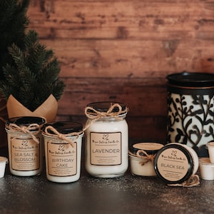 May include: An assortment of scented candles in glass jars with labels. The candles are arranged on a dark grey surface with a small Christmas tree in the background. The candle scents include Sea Salt & Blossom, Birthday Cake, Lavender, and Black Sea.