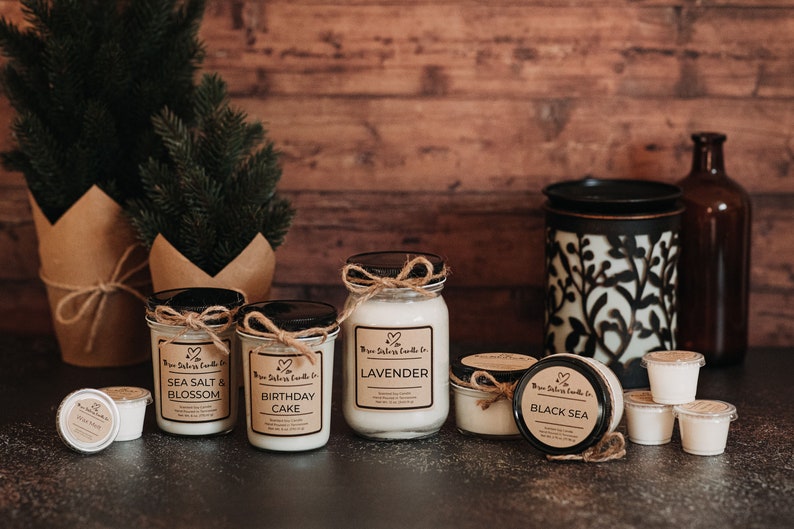 May include: An assortment of scented candles in glass jars with labels. The candles are arranged on a dark grey surface with a small Christmas tree in the background. The candle scents include Sea Salt & Blossom, Birthday Cake, Lavender, and Black Sea.