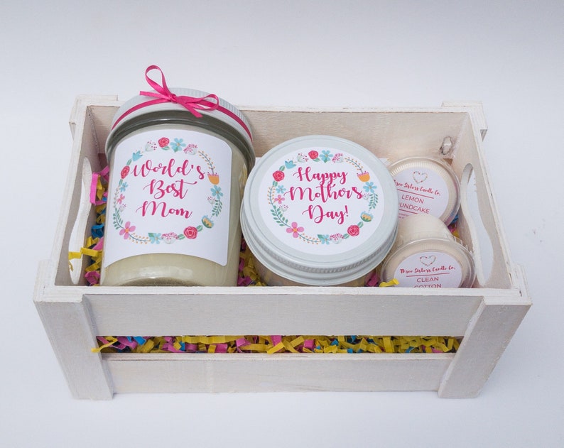 Op de afbeelding: Een witte houten kist gevuld met kleurrijk tissuepapier en drie kaarsen. De kaarsen zitten in glazen potten met etiketten waarop staat "World's Best Mom", "Happy Mother's Day!" en "Lemon Poundcake" en "Clean Cotton".
