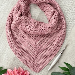 May include: A pink crocheted triangle scarf with a textured stitch pattern. The scarf is laid flat on a light wood surface with a crochet hook beside it.