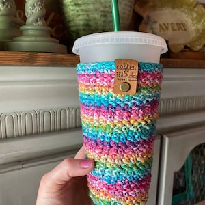 Könnte beinhalten: Ein regenbogenfarbener Häkelbecherwärmer mit einem braunen Lederanhänger, auf dem "coffee teach repeat" steht.