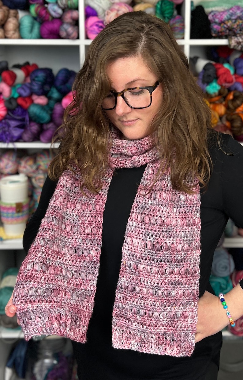 May include: A pink and white crocheted scarf with a textured pattern. The scarf is being held by a person wearing a black shirt.