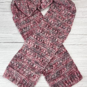 May include: A pink and grey crocheted scarf with a textured pattern. The scarf is folded in half and laid flat on a white wooden surface.