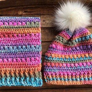 May include: A colourful crocheted hat and matching scarf with a white pom pom on the hat. The hat and scarf are made with a variety of colours, including pink, purple, blue, and orange.