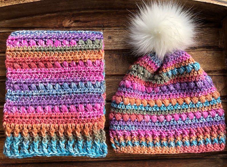 May include: A colourful crocheted hat and matching scarf with a white pom pom on the hat. The hat and scarf are made with a variety of colours, including pink, purple, blue, and orange.