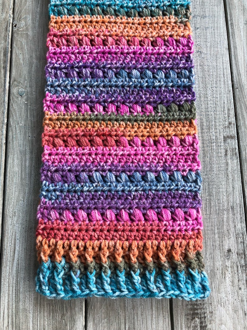 May include: A colourful crocheted scarf with stripes of teal, orange, pink, purple, and brown. The scarf has a textured stitch pattern with small bumps throughout.