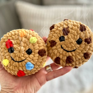 May include: Two crocheted cookie plush toys with smiling faces. One cookie is brown with chocolate chips and the other is tan with colourful sprinkles.
