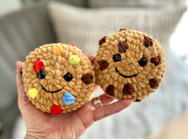 May include: Two crocheted cookie plush toys with smiling faces. One cookie is brown with chocolate chips and the other is tan with colourful sprinkles.