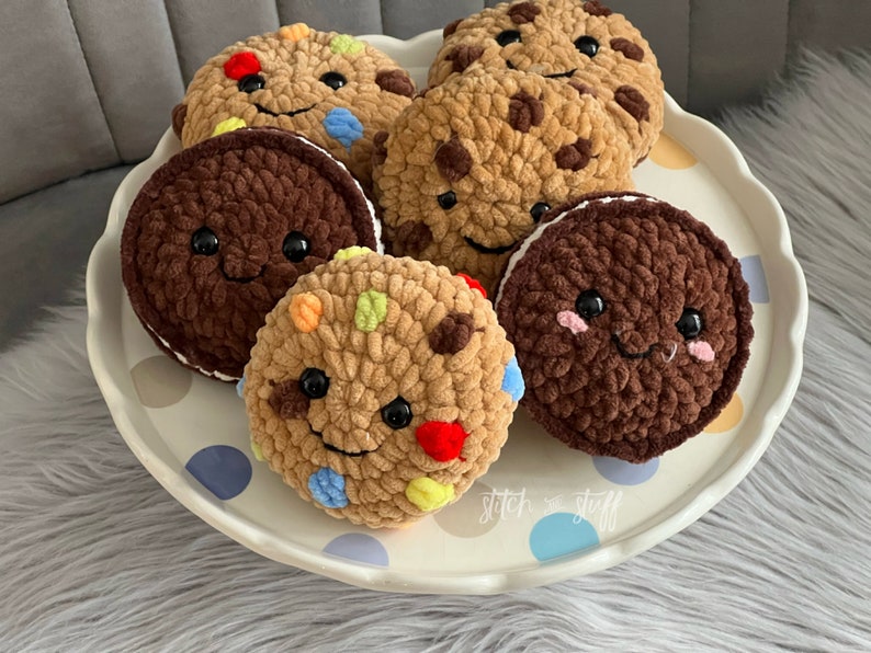 May include: Five crocheted cookies with smiling faces on a white plate with blue and brown polka dots. The cookies are brown, tan, and a mix of brown and colourful sprinkles. The plate has the text "stitch & stuff" on it.