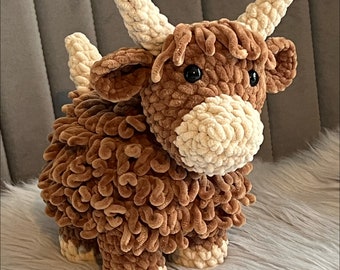 May include: A brown and white crocheted stuffed animal resembling a Highland cow. The cow has a fluffy brown body, white horns, and a white nose.