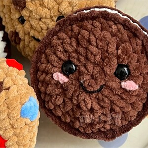May include: A brown plush cookie with a white frosting rim and a smiling face. The cookie has black eyes and pink cheeks. The cookie is sitting on a white surface with colourful polka dots.