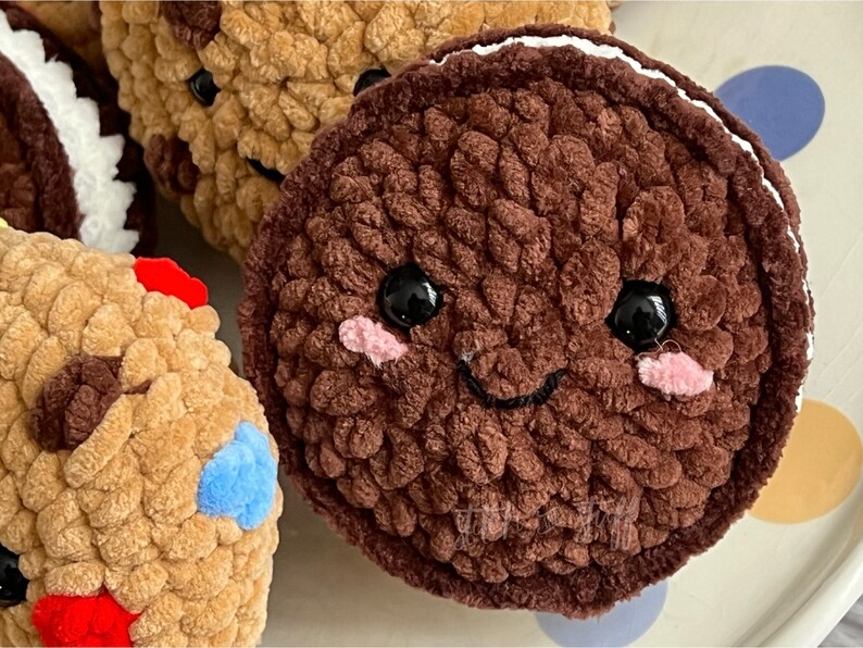 May include: A brown plush cookie with a white frosting rim and a smiling face. The cookie has black eyes and pink cheeks. The cookie is sitting on a white surface with colourful polka dots.