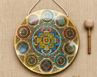 May include: A circular brass gong featuring a central square mandala design surrounded by eight smaller circular mandalas in various colors including red, blue, green, and yellow. A wooden mallet with a textured head is positioned to the right.