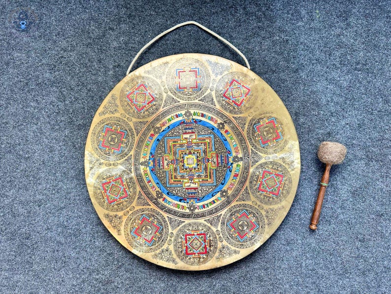 May include: A circular brass gong featuring an intricate, colorful mandala design at its center and eight smaller mandala patterns around the perimeter. The gong includes a metal hanging loop at the top and a wooden mallet with a textured head.