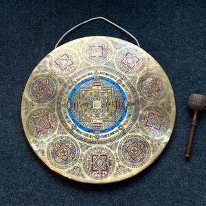 May include: A circular brass gong featuring an intricate, colorful mandala design in the center and eight smaller mandala patterns around the perimeter. The gong includes a white hanging cord and a wooden mallet with a textured, rounded head.