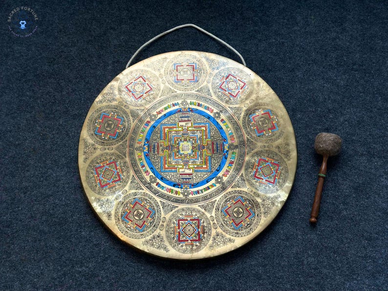May include: A circular brass gong featuring an intricate, colorful mandala design in the center and eight smaller mandala patterns around the perimeter. The gong includes a white hanging cord and a wooden mallet with a textured, rounded head.