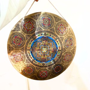 May include: A circular brass gong featuring an intricate central mandala design surrounded by eight smaller geometric patterns. The metal surface is engraved with detailed motifs and accented with vibrant blue, red, and green enamel colors.