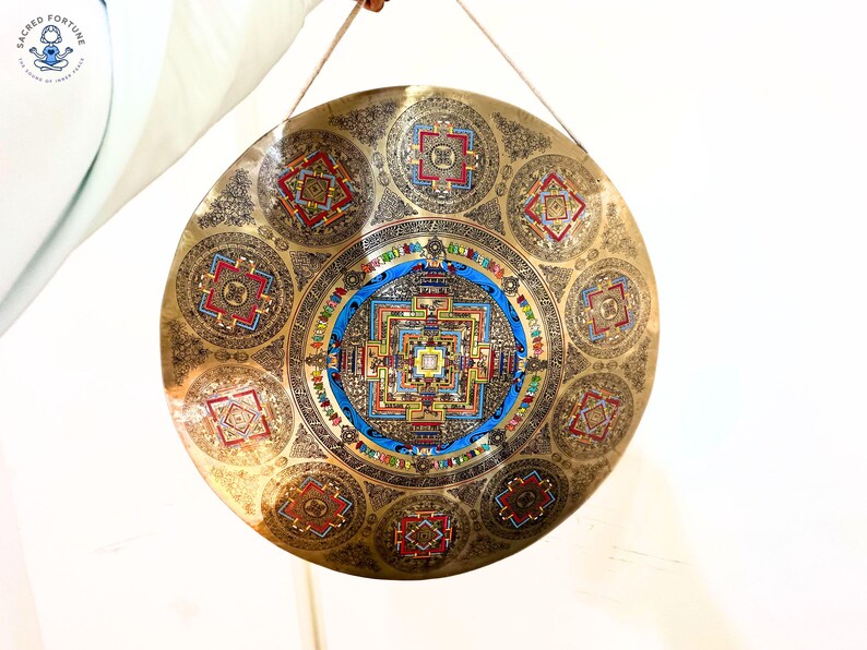 May include: A circular brass gong featuring an intricate central mandala design surrounded by eight smaller geometric patterns. The metal surface is engraved with detailed motifs and accented with vibrant blue, red, and green enamel colors.