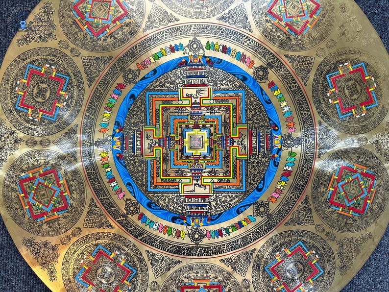 May include: A circular brass plate featuring an intricate mandala design with a central square structure, surrounded by concentric rings of blue, black, and gold patterns. Smaller square motifs are arranged around the outer edge.