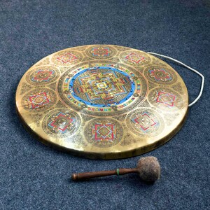 May include: A circular brass gong featuring an intricate, colorful mandala design at its center and eight smaller geometric patterns around the perimeter. A white cord is attached to the rim, and a wooden mallet with a felt head rests below the gong.