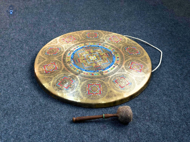 May include: A circular brass gong featuring an intricate, colorful mandala design at its center and eight smaller geometric patterns around the perimeter. A white cord is attached to the rim, and a wooden mallet with a felt head rests below the gong.