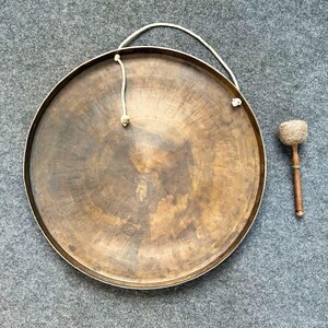May include: A large, circular bronze gong with a weathered, dark patina and a raised central boss. A thick, off-white rope is threaded through two holes at the top for suspension. Beside the gong lies a wooden mallet with a padded, textured head.