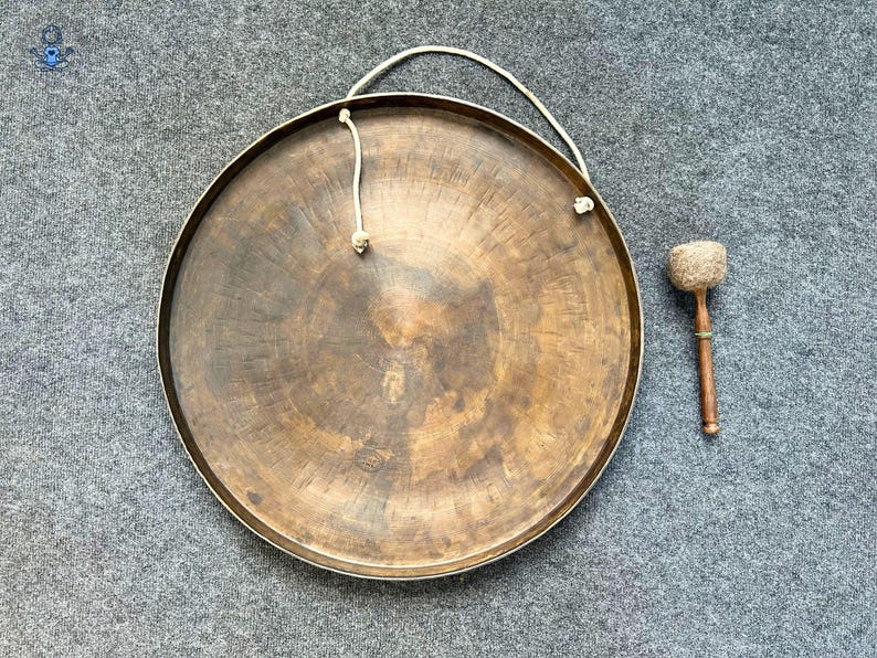 May include: A large, circular bronze gong with a weathered, dark patina and a raised central boss. A thick, off-white rope is threaded through two holes at the top for suspension. Beside the gong lies a wooden mallet with a padded, textured head.